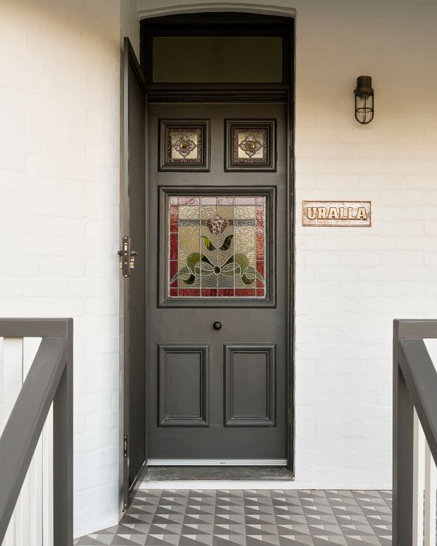 Front entry porch featuring a dark grey, traditionally detailed timber door with decorative stained-glass panels and raised moulded panels. The door is set in a white-painted brick facade, with a vintage-style wall light and a ceramic address plaque reading “URALLA.” A geometric monochrome tiled floor and dark timber porch railings complete the welcoming heritage-inspired entrance.