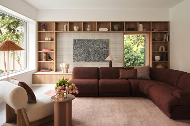 A warm, refined living room with a large brown sectional sofa, light upholstered armchair, round side table, floor lamp, and soft pink-beige rug. A full-width built-in timber shelving and cabinetry wall frames a tiled feature panel and artwork, with large windows bringing in greenery and natural light.