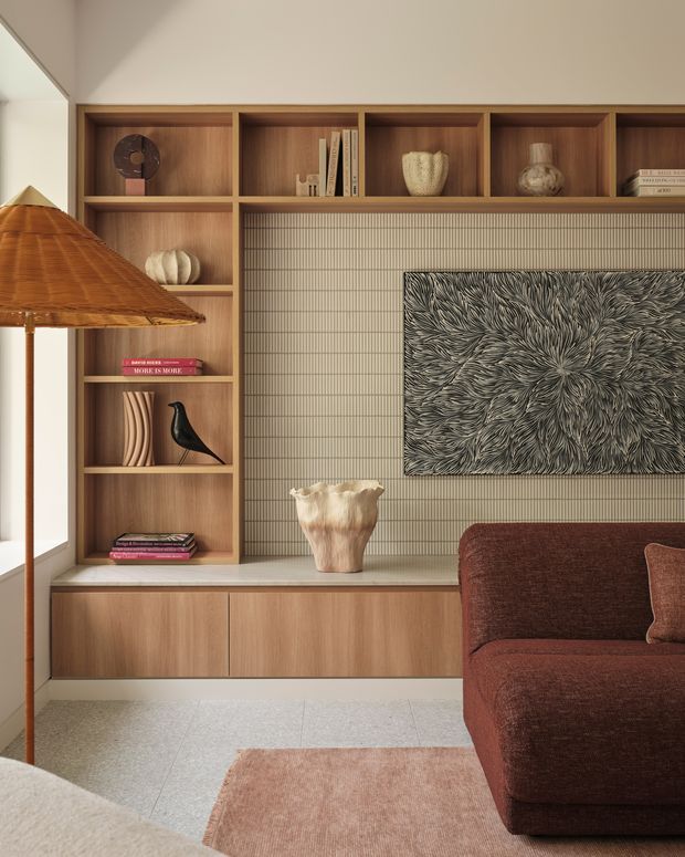 A warm, refined living room vignette featuring a custom timber built-in shelving and low cabinetry wall. The joinery incorporates open display niches, books, ceramics and sculptural objects, with a vertically lined neutral feature surface behind a large monochrome artwork. A rust-brown upholstered sofa, woven pendant light, pale stone-toned floor and muted rug create a tactile, earthy interior.