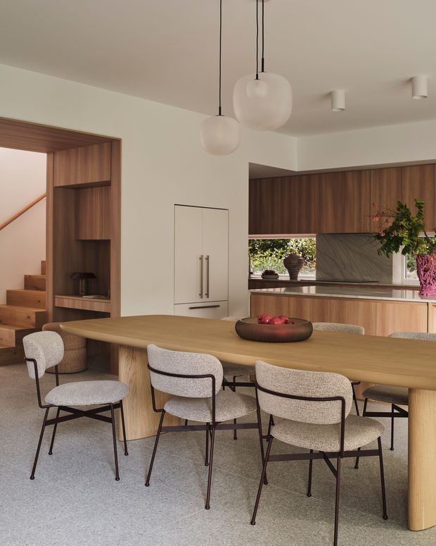 A refined open-plan dining and kitchen interior with a long rounded timber dining table surrounded by upholstered dining chairs. Warm timber cabinetry and wall panelling frame the kitchen, with a pale stone or marble-look benchtop and splashback, integrated white appliances, and sculptural pendant lights. A timber-lined stair and built-in shelving are visible to the left, while soft neutral finishes create a calm contemporary atmosphere.