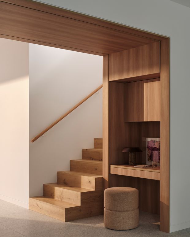 A warm, refined interior view of a timber-lined stair and integrated built-in joinery. The staircase has broad solid-wood treads and a matching handrail against pale plaster walls. Beside it is a full-height dark timber feature unit with open shelving, concealed cabinetry, a small desk surface, and a round upholstered stool. The space is softly lit with a neutral palette and minimalist detailing.