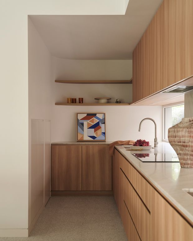 A compact, warm-toned contemporary kitchen with extensive vertical-grain timber cabinetry, integrated upper storage, open timber shelves, and a pale stone benchtop. A curved chrome faucet, undermount sink, woven basket, fruit bowl, and colourful geometric artwork add detail. The kitchen has a narrow galley-like layout, soft neutral walls, and a partially recessed ceiling creating subtle architectural depth.