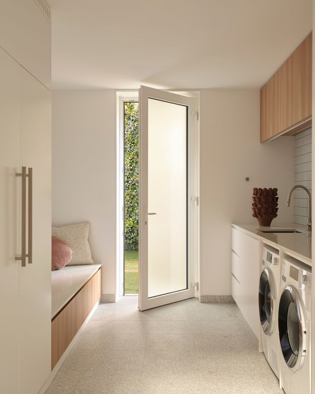 A bright, compact contemporary laundry room with a stacked-height washer and dryer beneath a pale stone benchtop, white cabinetry, and a light horizontal tiled splashback. Warm timber cabinetry and a built-in cushioned bench seat line the opposite wall. A full-height glazed door opens to greenery outside, bringing in natural light. The room has light neutral finishes and a practical, refined fit-out.