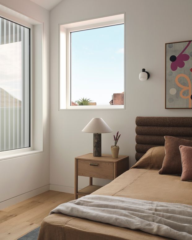 A bright, warm bedroom corner with a low platform bed, textured rounded brown upholstered headboard, tan bedding, and layered pillows. A timber bedside table holds a sculptural lamp and small vase, while large white-framed windows provide abundant natural light. Neutral walls, timber flooring, a wall sconce, and colourful abstract artwork complete the calm contemporary interior.