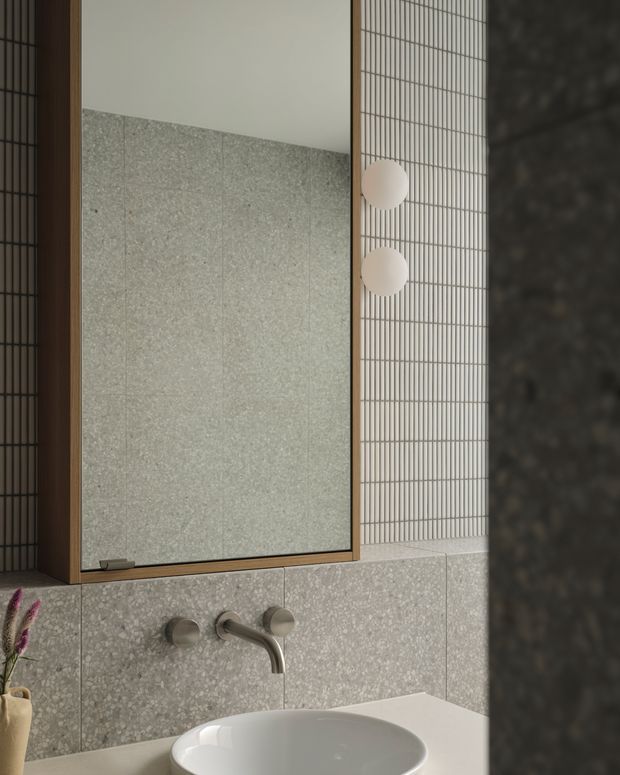 Close-up view of a refined contemporary bathroom vanity area featuring a round white vessel basin, wall-mounted brushed metal tapware, a large timber-framed mirror, and pale grey terrazzo or stone wall cladding. A vertical grid-patterned tiled wall with spherical wall lights adds texture and detail.