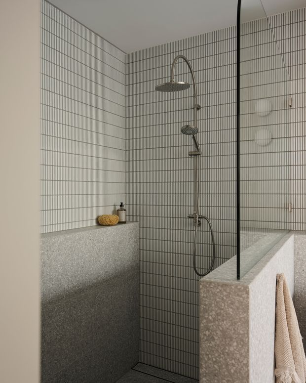 A minimalist bathroom shower area with fine vertically lined light grey wall tiles, a dark metal-framed glass shower screen, brushed metal rain shower and handheld fixture, and terrazzo-style partition walls and ledges. Neutral warm-grey tones and understated fixtures create a refined, contemporary atmosphere.