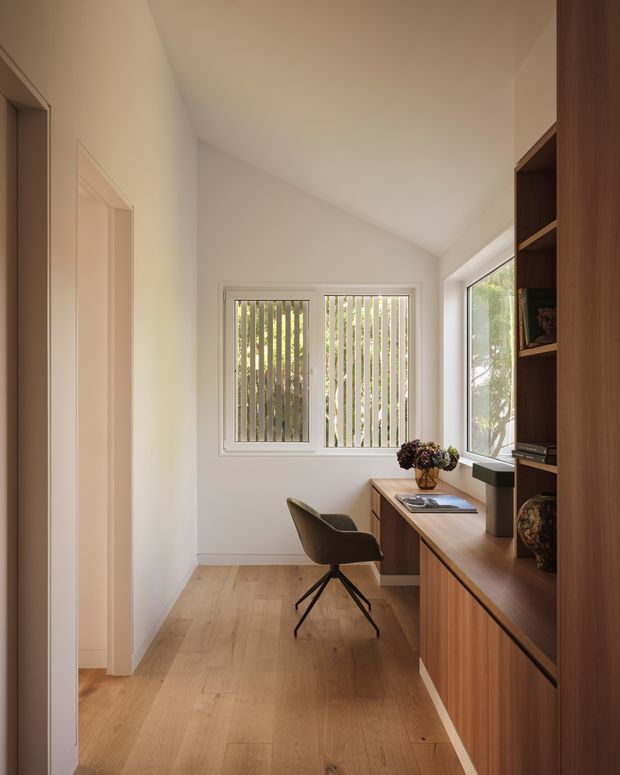 A compact, light-filled home office with a built-in timber desk and floor-to-ceiling shelving along one wall. A modern upholstered swivel chair faces the desk, while a pair of windows with vertical blinds provide filtered natural light. The room has white walls, a warm timber floor, and a clean, restrained contemporary fit-out.