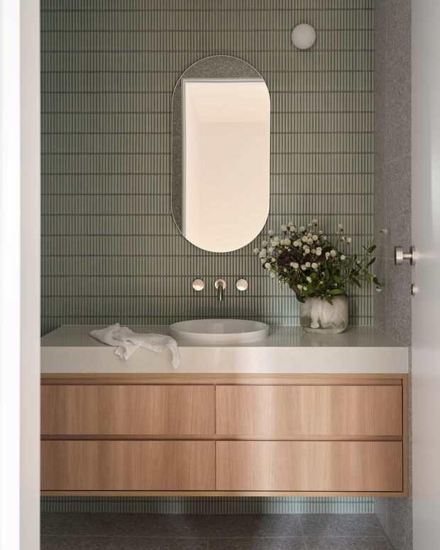 A refined bathroom vanity viewed straight on, featuring a wall-mounted timber vanity with four drawers, a pale solid-surface benchtop, round vessel basin, brushed metal wall-mounted tapware, and an arched illuminated mirror. The walls and floor are finished in dark green-grey vertically ribbed tiles, with a sculptural wall light and floral arrangement adding softness.