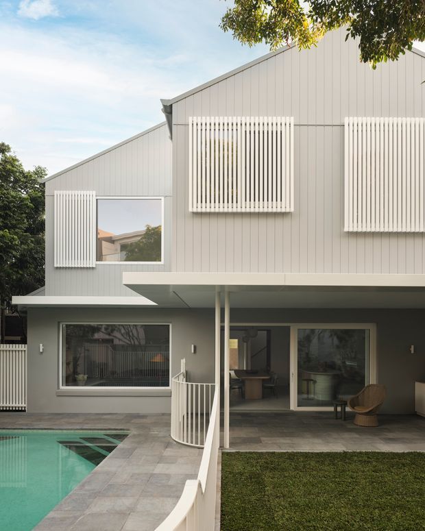 Contemporary two-storey residential home with a pale grey vertically clad upper level, projecting flat-roofed lower terrace, and distinctive white vertical screening over windows. The view looks across a paved pool terrace and lawn toward the covered outdoor living area, with interior dining and seating visible through the glazing.