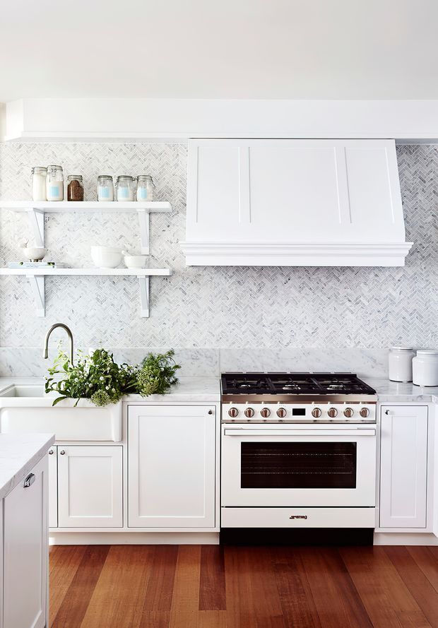 Bright, refined white kitchen with a freestanding stainless-steel and white range cooker, white Shaker-style cabinetry, open shelving, a marble-look benchtop, and a pale herringbone tile splashback. A large farmhouse-style sink with a brass-toned tap sits beside fresh herbs, while warm timber flooring adds contrast.
