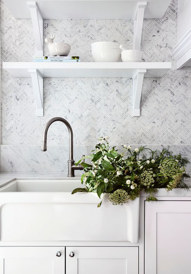 Bright, close-up view of a farmhouse-style kitchen sink area. A white apron-front sink sits beneath a tall brushed metal gooseneck faucet, with a marble backsplash ledge and pale grey-white herringbone tile backsplash. White built-in cabinetry and open bracketed shelving display ceramic bowls and vases, while a leafy floral arrangement adds natural greenery. The palette is predominantly white and light grey with subtle warm metallic accents.