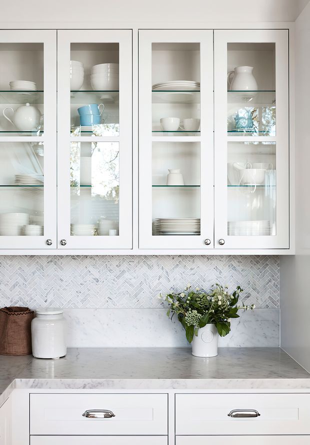 A bright, refined kitchen cabinetry detail featuring white framed glass-front upper cabinets displaying white crockery and glassware, a pale marble benchtop and backsplash, and a light grey-white herringbone tile splashback. White shaker-style drawers with polished metal pulls appear below, with a vase of greenery and woven container adding subtle natural accents.