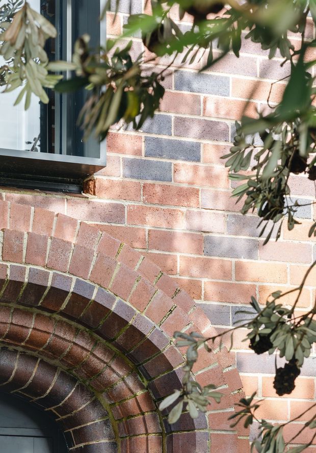 Close-up exterior detail of a red and muted blue-grey brick facade, featuring a large arched brick opening with radial voussoirs and a dark-framed window above. Sunlit leafy branches partially obscure the masonry, highlighting the textured brickwork and weathered character.