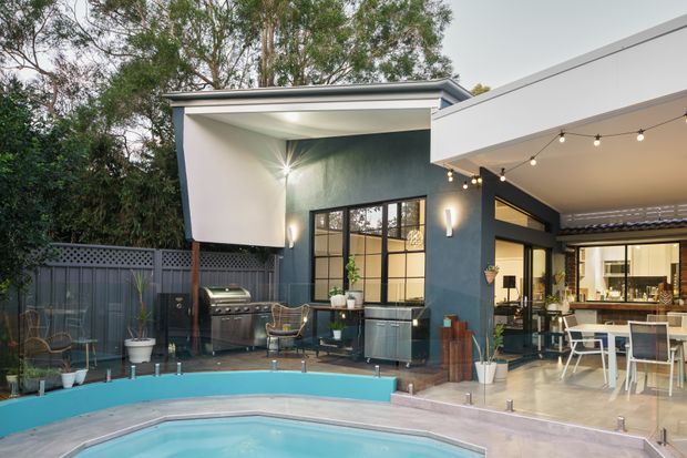 Contemporary residential outdoor entertaining area viewed from beside a swimming pool, with a covered patio, outdoor dining furniture, barbecue, string lights, large glazed openings, and a dark feature wall. The space opens onto a landscaped backyard surrounded by trees and fencing.