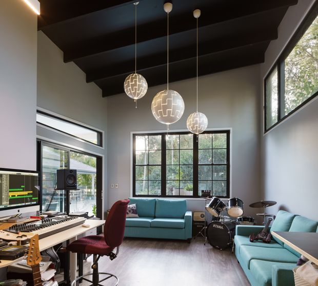 A bright, high-ceilinged music studio and lounge with exposed dark timber ceiling beams, large black-framed windows, a computer and keyboard workstation, drum kit, guitars, pendant globe lights, and teal sofas with a burgundy office chair. The room has a creative, contemporary studio atmosphere with views to greenery outside.