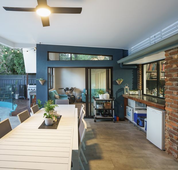 A bright open-plan dining and entertaining area with a long white dining table, chairs, indoor plants, and a ceiling fan. The room includes a dark blue feature wall, large glazed doors opening toward a music or sitting area, exposed brickwork, timber shelving, and a compact kitchen or bar zone with cabinetry and appliances. A covered outdoor patio, barbecue, and pool are visible beyond the glazing.