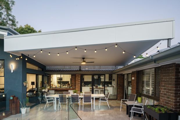 A contemporary covered outdoor courtyard entertaining area adjoining a brick house. The space features a large flat-roofed canopy, exposed brick walls, extensive glazing and sliding doors, a tiled or polished concrete floor, outdoor dining furniture, built-in planter boxes, ceiling fans, and festoon lighting. The courtyard appears to connect directly with the interior kitchen and living spaces.
