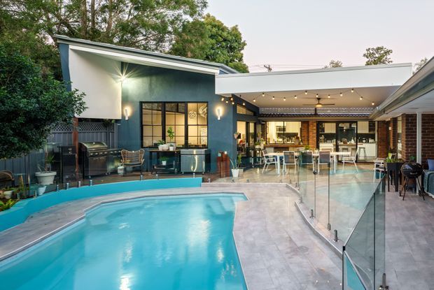 A renovated courtyard-style outdoor entertaining area with a turquoise swimming pool in the foreground, surrounded by pale tiled paving and frameless glass pool fencing. The space includes a covered outdoor kitchen and dining area with pendant-style string lighting, bar seating, built-in cabinetry, and a barbecue. Dark teal and white walls contrast with exposed brick detailing, while established trees and planting frame the courtyard.