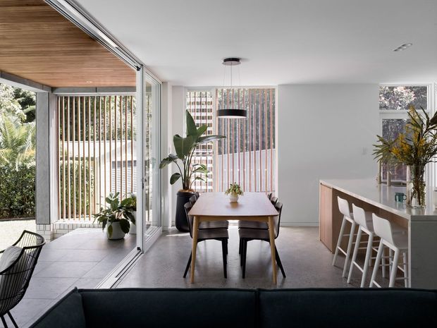 Bright open-plan contemporary interior combining a dining area, kitchen island and living space. Full-height glazed sliding doors open to a leafy exterior, with vertical timber screening, a timber-lined ceiling over the terrace, pale walls, concrete-look flooring, and restrained modern furniture. A dining table sits centrally beneath a black pendant light, while a pale kitchen island with white stools occupies the right side.