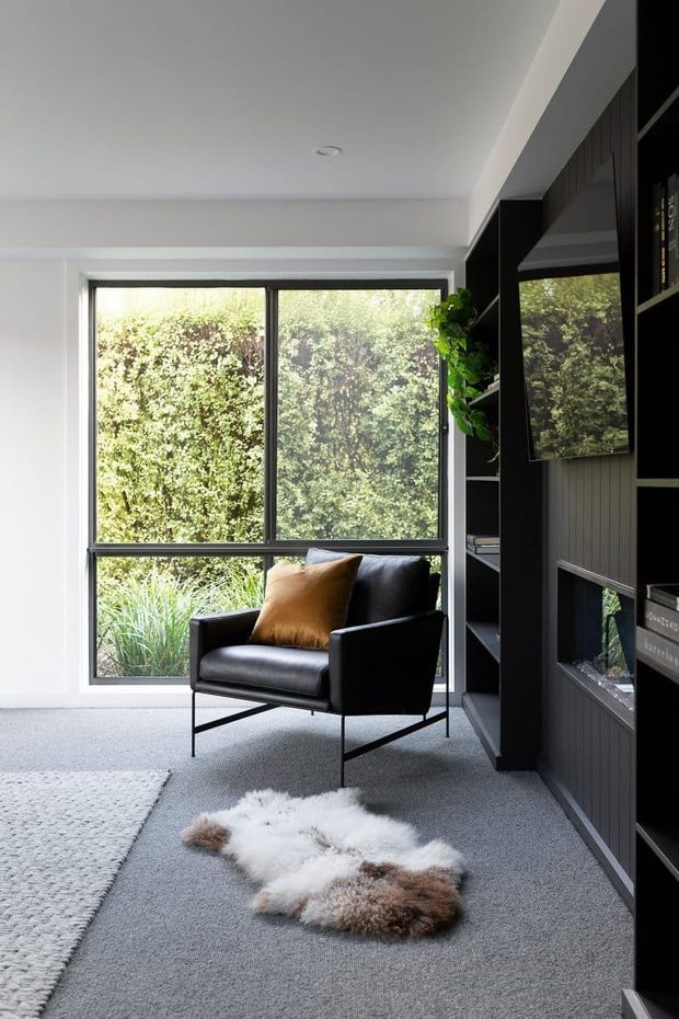 A refined contemporary living room or reading nook with a black leather armchair positioned beside full-height garden glazing. The room features dark built-in shelving and media cabinetry with vertical wall panelling, a grey carpeted floor, a textured light rug, and a small sheepskin accent rug. Abundant greenery is visible outside the window, creating a calm connection to the garden.