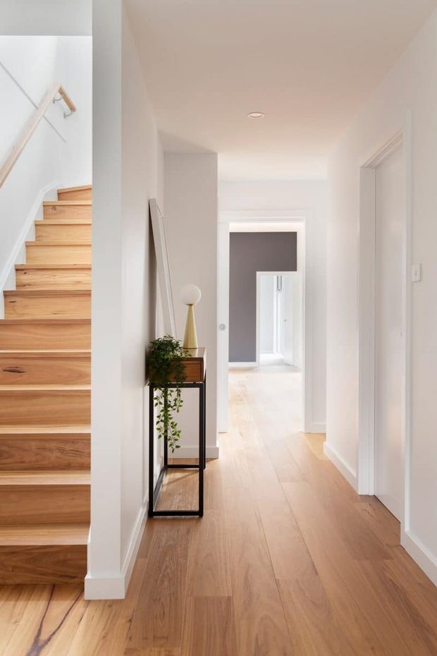 A bright, narrow residential hallway with warm timber flooring, white walls and trim, and a timber staircase rising along the left side. A slim black console table with a small plant and lamp sits against the wall, while multiple doorways and a softly contrasting grey wall lead further into the home. The space has clean, understated contemporary detailing and abundant natural light.