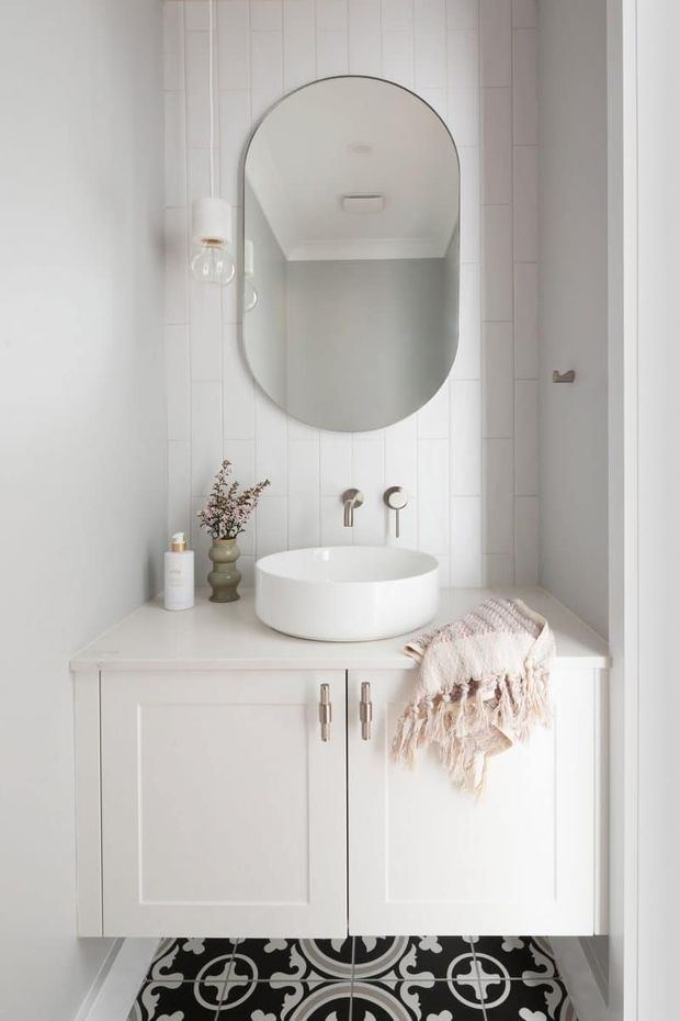 A compact, bright bathroom vanity with a white vessel basin on a pale countertop, brushed metal wall-mounted tapware, and a large rounded-rectangle mirror. White vertical wall tiles and white panelled cabinetry create a clean, refined look, while patterned black-and-white floor tiles and a soft fringed towel add contrast and texture.