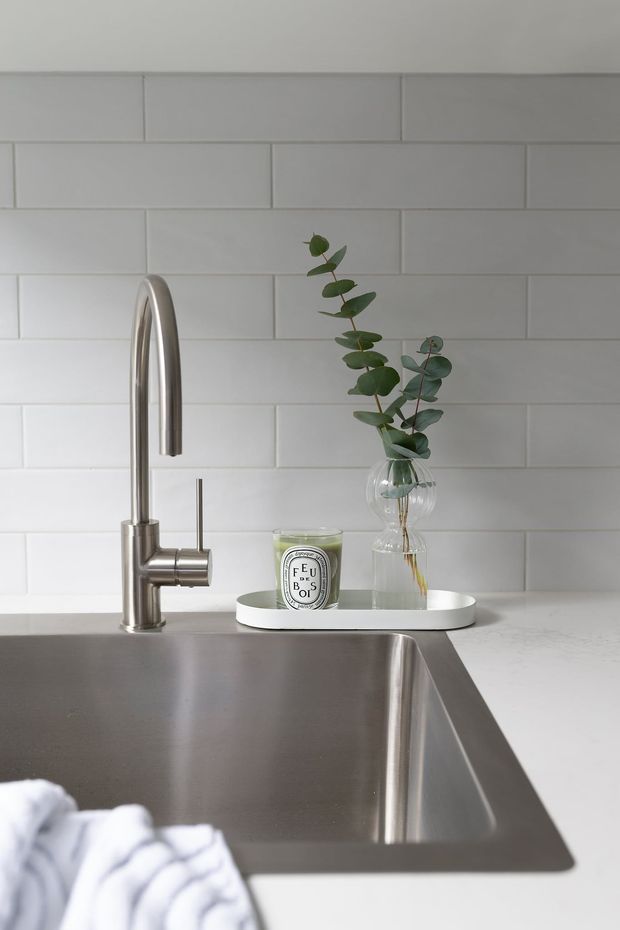 Close-up of a contemporary kitchen sink area featuring a brushed stainless-steel gooseneck faucet, deep integrated stainless-steel sink, pale engineered stone countertop, and white horizontal subway-tile splashback. A small tray with a candle and glass vase of eucalyptus provides subtle styling.