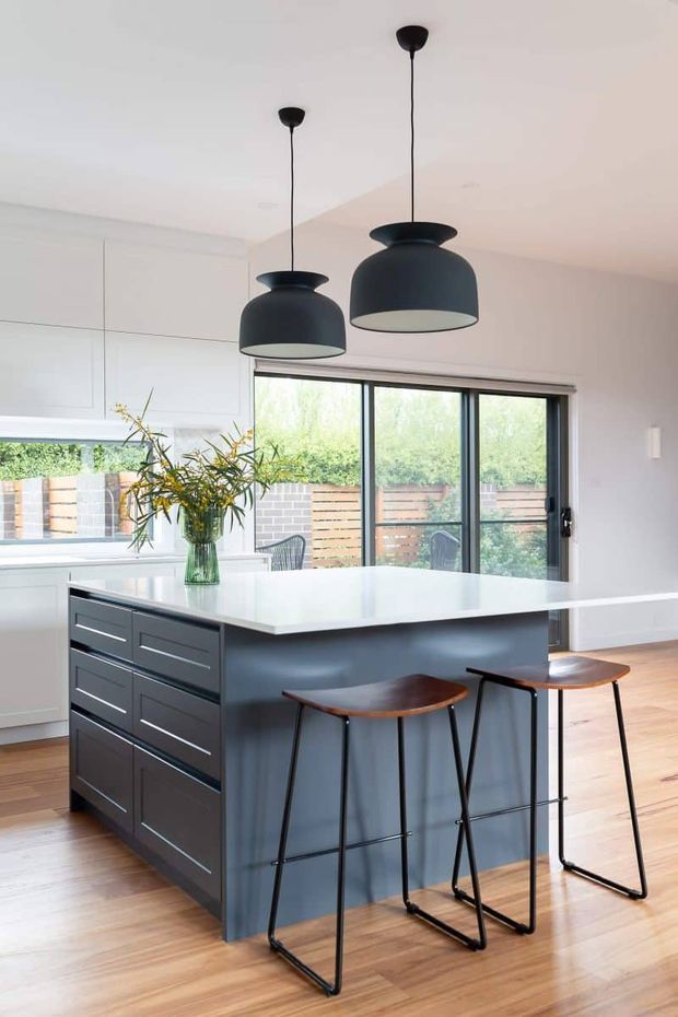 Bright contemporary kitchen featuring a large dark island with a white benchtop, integrated drawers, two timber-and-metal bar stools, pale timber flooring, white built-in cabinetry, and oversized matte-black pendant lights. Full-height glazed doors open toward a planted outdoor area, bringing in abundant natural light.