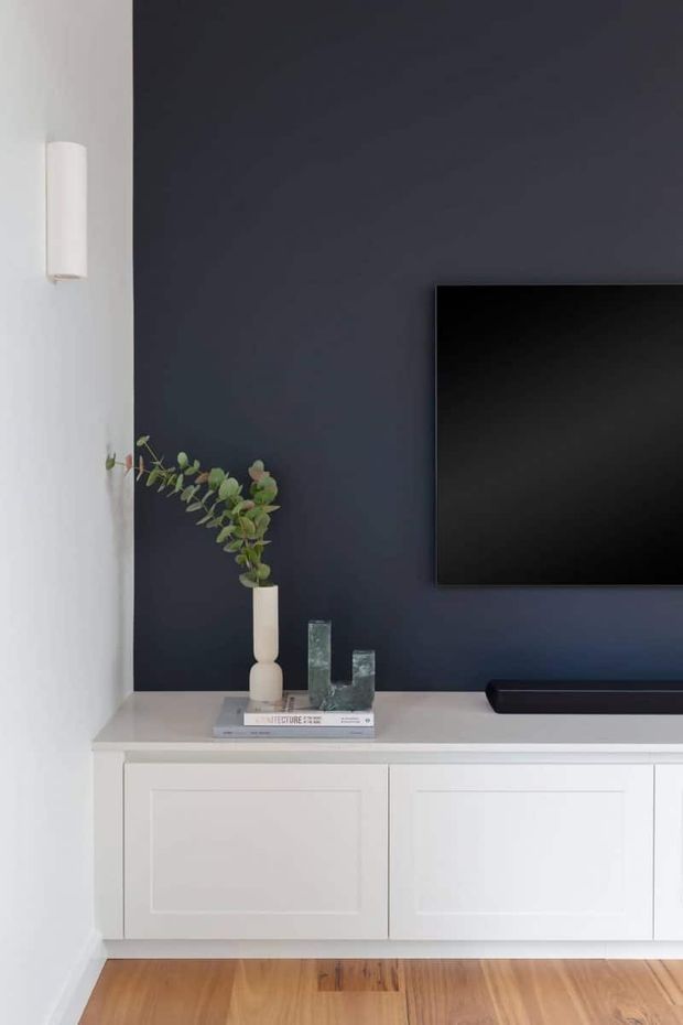 A minimalist media wall featuring a large black television mounted on a deep charcoal-blue accent wall, with a long white paneled built-in media cabinet below. Simple decorative objects and a vase with greenery sit on the cabinet, while warm timber flooring contrasts with the cool dark wall. A slim white wall sconce is mounted on the adjacent white wall.