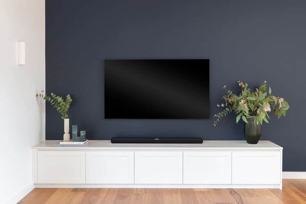 A clean, minimalist living room media wall featuring a wall-mounted black television on a deep navy-blue accent wall, with a long white paneled built-in entertainment cabinet below. Decorative vases with leafy branches sit at both ends, and warm timber flooring contrasts with the cool wall colour.