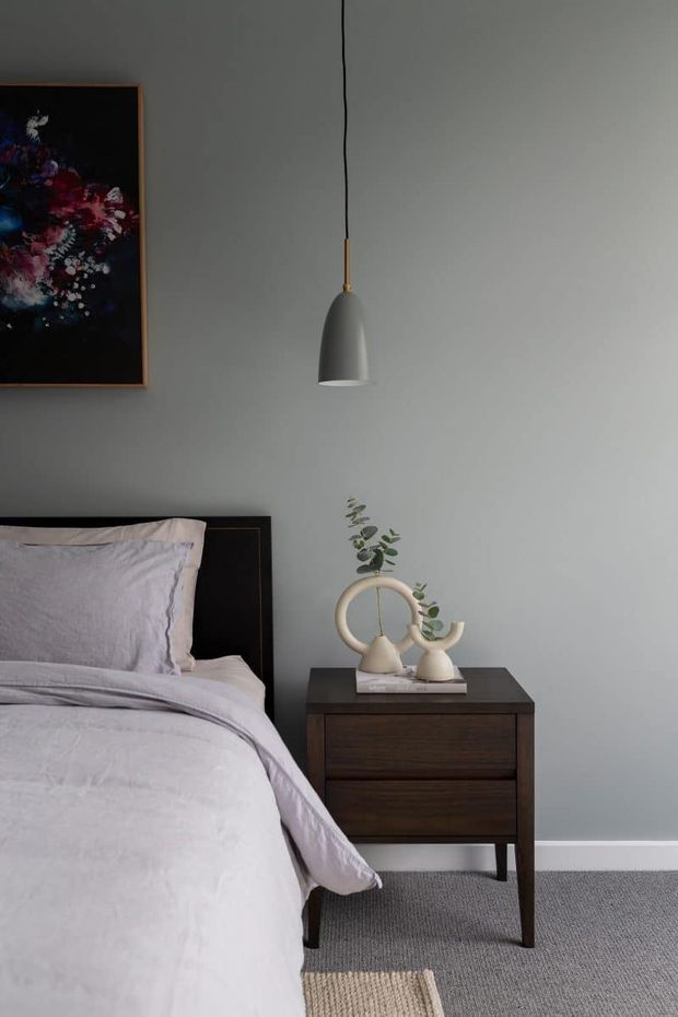 A softly lit bedroom vignette featuring a neatly made bed with pale grey bedding, a dark timber bedside table, a minimalist pendant light, framed floral artwork, and small sculptural ceramic vases with greenery. The room has a calm, understated contemporary feel with grey walls and carpet.