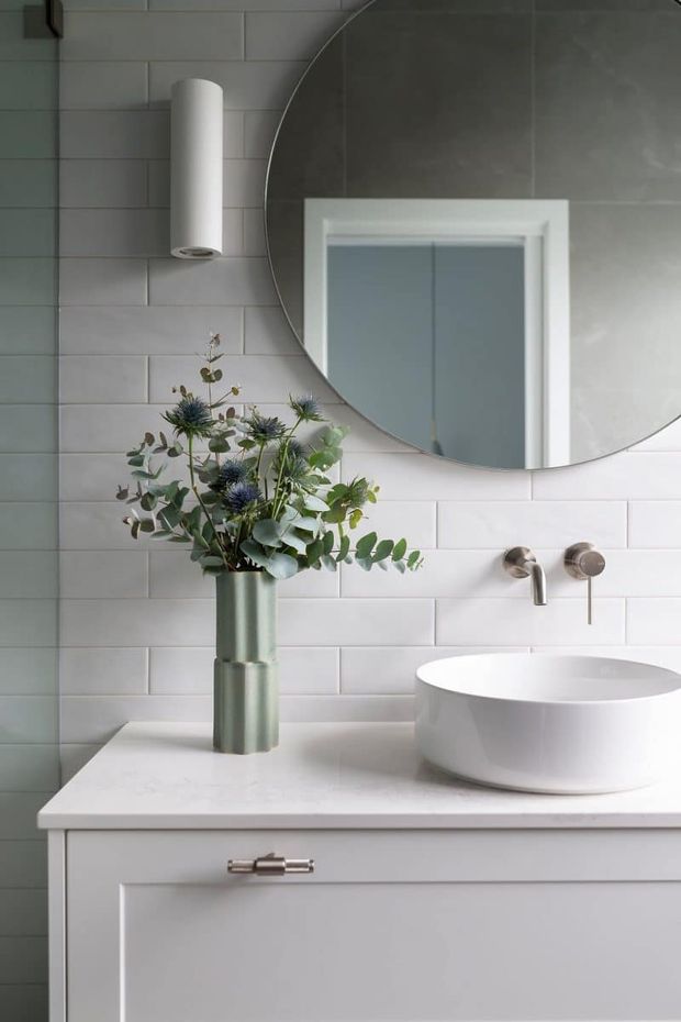 A close-up of a refined bathroom vanity with white rectangular wall tiles, a white vessel basin on a white benchtop, a pale grey-white vanity with framed cabinetry, brushed metal wall-mounted tapware, and a large round mirror. A green botanical arrangement in a metallic vase adds natural contrast.