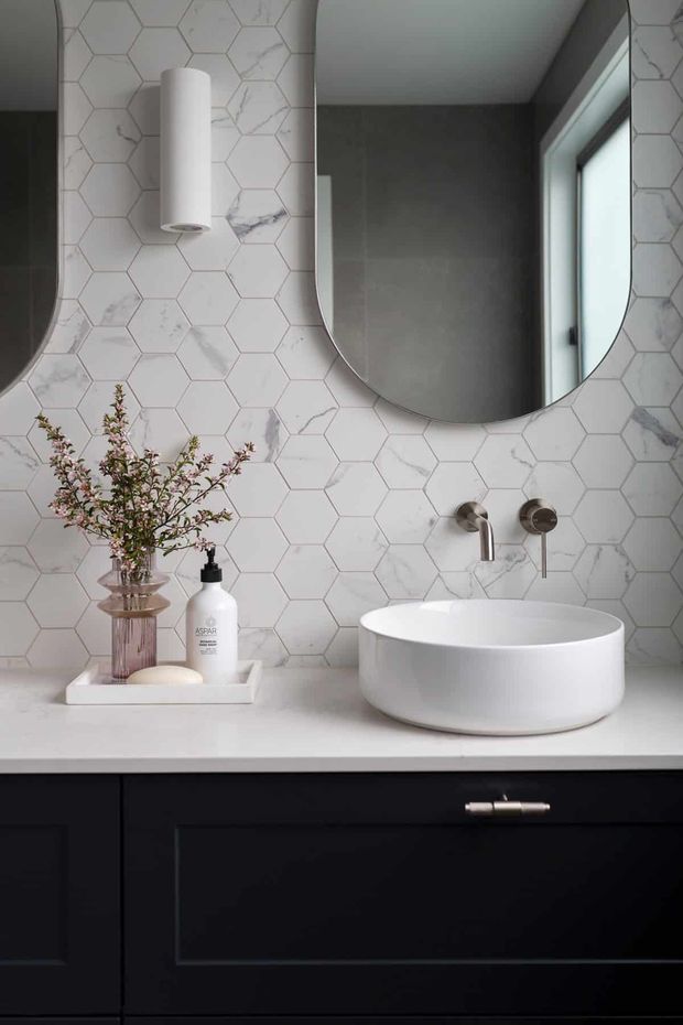 A refined bathroom vanity featuring a white vessel basin on a white benchtop, dark navy or black shaker-style cabinetry, and a white marble-look hexagonal tile splashback. A rounded rectangular mirror, wall-mounted cylindrical light, brushed metal tapware, and a small decorative tray complete the sophisticated monochromatic scheme.