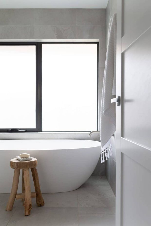 A bright, minimalist bathroom featuring a freestanding white bathtub beneath a large black-framed window. Pale grey stone-look wall and floor tiles, white painted wall panelling, and a simple timber stool with a folded white towel create a clean, calm atmosphere.