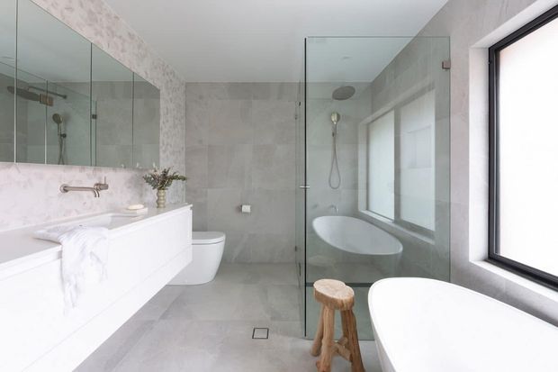 Bright contemporary bathroom featuring a freestanding white bathtub beside a large black-framed window, a frameless glass shower enclosure, wall-mounted white vanity, toilet, extensive mirrored surfaces, and pale grey stone-look wall and floor finishes.