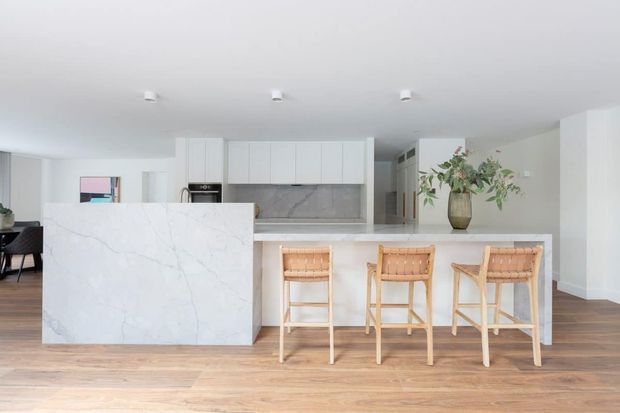Bright open-plan kitchen with a large white marble-look island, white flat-panel cabinetry, integrated stone splashback, and three woven timber bar stools. Warm timber flooring contrasts with the predominantly white interior, while a dining area and decorative greenery are visible in the background.