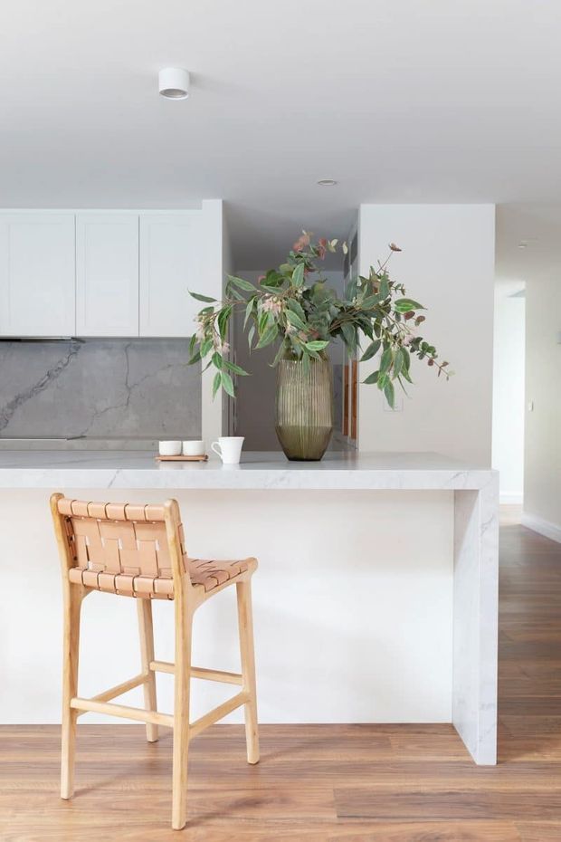 Bright, minimalist kitchen interior featuring a white stone island with a waterfall edge, white flat-panel cabinetry, a grey-veined stone splashback, timber flooring, and a single woven timber bar stool. A large vase of greenery adds a natural focal point. The kitchen opens toward a hallway or adjoining living area.