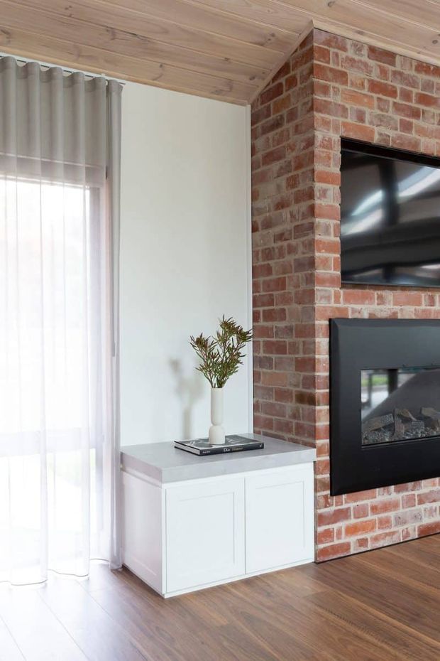 A bright living room corner featuring a red exposed-brick feature wall with a built-in black fireplace and wall-mounted television. A white built-in cabinet with a pale stone-like top sits beside sheer curtains, while the room has warm timber flooring and a timber-lined ceiling.