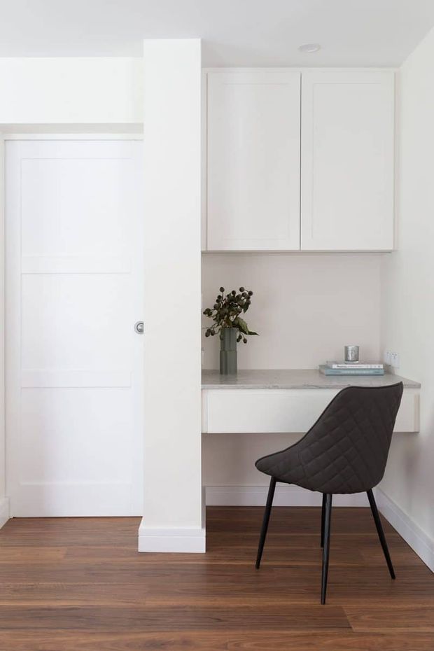 Bright, compact built-in study nook beside a white paneled interior door. The nook features white overhead cabinetry, a pale stone-look desktop, a small vase with dried foliage, and a dark quilted chair. Warm timber flooring and white walls create a clean, understated workspace.