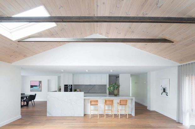 Bright open-plan kitchen and living area with a white marble-look island, white cabinetry, timber flooring, and a dramatic pitched timber ceiling with exposed dark beams and skylights. A dining area is visible to the left, while full-height curtains and glazing appear on the right.