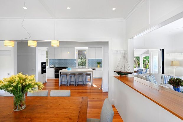 Bright open-plan coastal-style interior showing a dining area in the foreground, a kitchen with a central island and stools beyond, and a living room visible through a wide opening on the right. The space features white walls and detailed ceiling trim, warm timber flooring, white built-in cabinetry, a blue mosaic-tile kitchen splashback, pendant lighting, and abundant daylight.