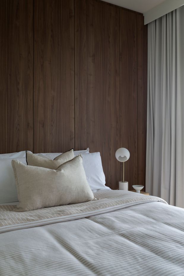A calm, softly lit bedroom featuring a bed with layered neutral linen bedding and cushions, a full-height dark timber wall behind the bed, a small spherical bedside lamp, and floor-to-ceiling sheer curtains. The room has a refined contemporary feel with warm natural materials and muted tones.