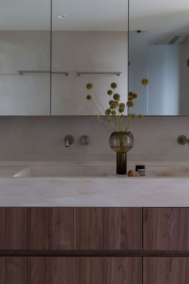 A refined contemporary bathroom vanity viewed straight on, featuring a large mirrored wall cabinet, pale stone or concrete-look benchtop with integrated rectangular basins, wall-mounted brushed metal tapware, and dark timber-grain cabinetry. A simple glass vase with dried botanical stems adds a muted decorative accent.