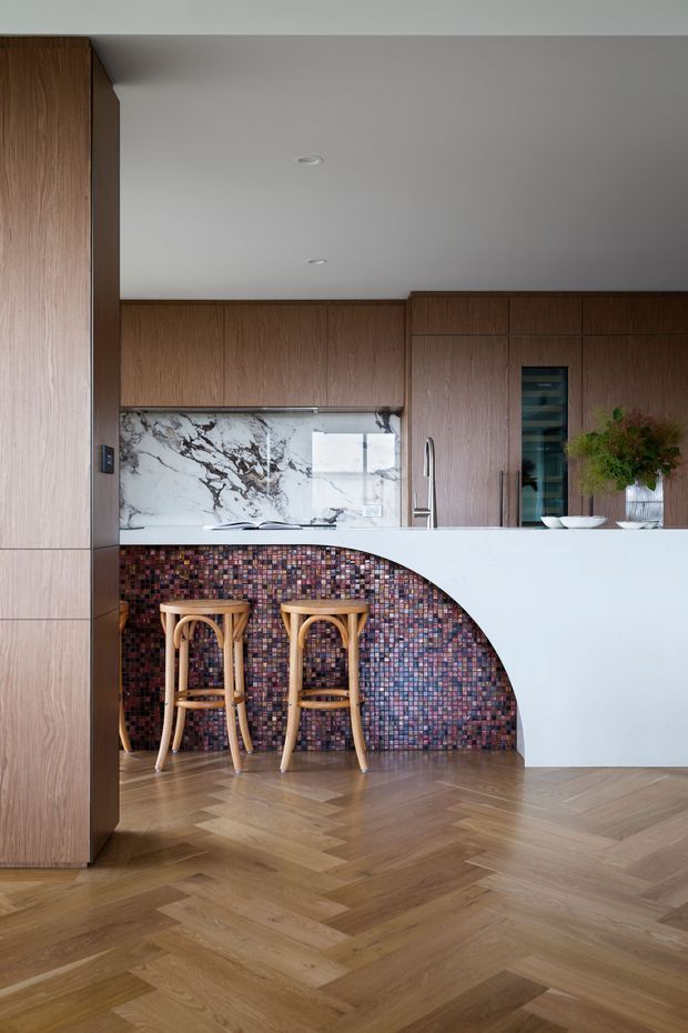 A refined contemporary kitchen featuring warm timber cabinetry, a white marble-look splashback, a white curved island or breakfast bar, and a richly patterned multicolour mosaic-tile feature beneath the counter. Two timber bar stools sit at the island. The room has a white ceiling with recessed downlights and a warm herringbone timber floor.