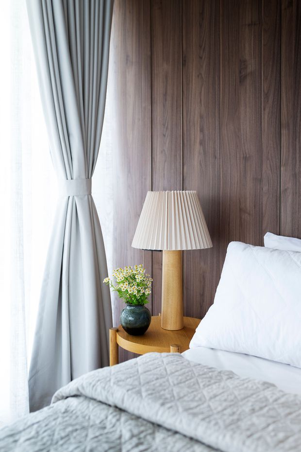 A softly lit bedroom corner featuring a bed with white pillows and a pale quilted cover, a warm timber bedside table, a pleated table lamp, a dark ceramic vase with flowers, full-length grey curtains, and a rich vertical timber-panelled wall behind the bed.