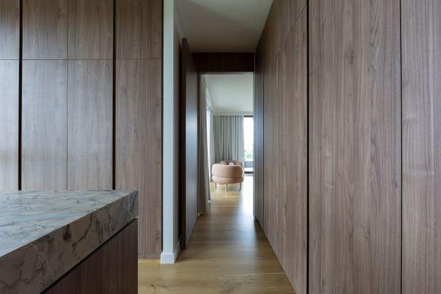 A narrow, refined interior passage lined on both sides with full-height warm timber wall panelling and integrated cabinetry. A marble-topped island or benchtop is visible in the foreground, while the corridor opens toward a softly furnished bedroom or living area with a rounded upholstered chair and light curtains. The space has timber flooring, concealed storage, and a calm, minimalist material palette.