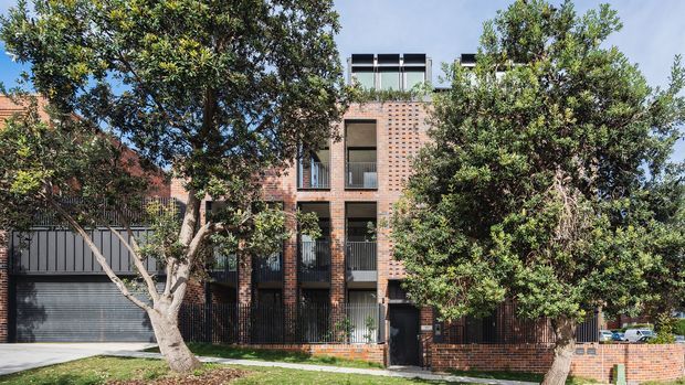 Exterior view of a contemporary multi-storey brick townhouse or apartment-style residence, partially screened by mature trees. The facade features warm red-brown brickwork, dark metal-framed windows and balconies, a large dark garage door, and a flat roofline with rooftop elements. A low brick boundary wall and landscaped verge are visible in front.