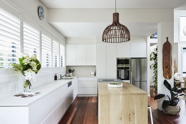 Bright, spacious contemporary kitchen with white flat-panel cabinetry, a large light timber island, dark timber flooring, white tiled backsplash, stainless-steel appliances, plantation shutters, and a sculptural woven pendant light. Indoor plants and natural materials add warmth to the predominantly white scheme.