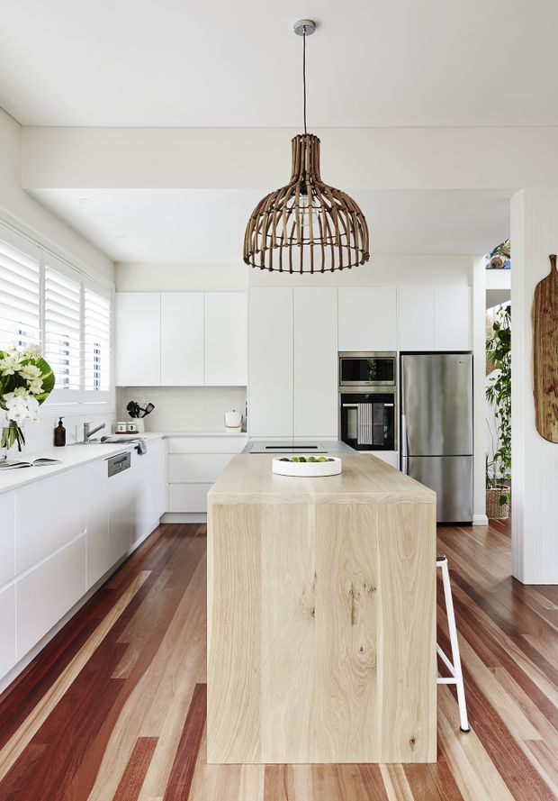 Bright contemporary kitchen with white flat-panel cabinetry, a large light timber island, stainless-steel appliances, and richly varied timber flooring. A woven rattan pendant light hangs above the island, while plantation shutters, indoor greenery, and timber accessories add warmth and texture.
