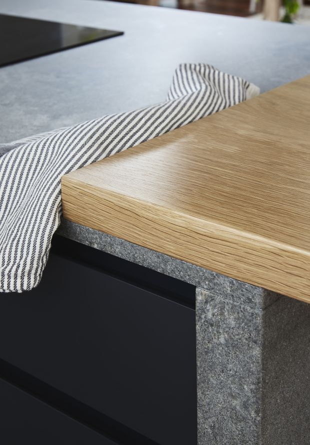 Close-up detail of a contemporary kitchen island showing a light oak-look benchtop with a rounded edge, dark matte cabinetry, and a grey textured stone side panel. A striped tea towel drapes over the island, with a black cooktop partially visible in the background.
